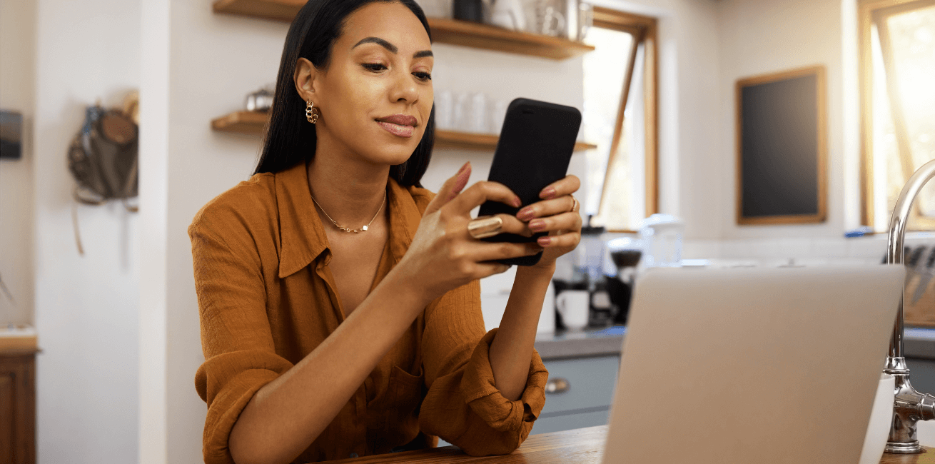 Young woman sitting in front of a laptop and using a smartphone to read an SMS from her financial services institution